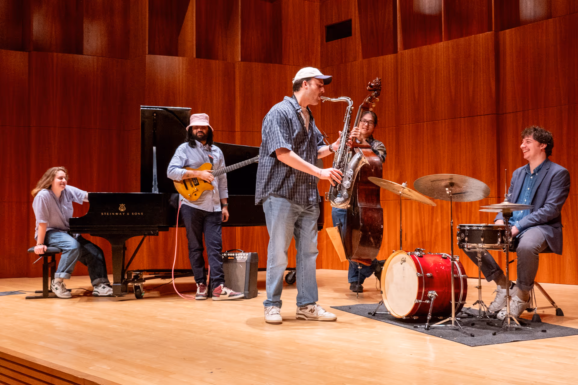 Eshaan Sood and the Sonic Alchemists perform with relaxed joy on a wooden stage. He stands near a grand piano and an upright bass, while a saxophonist and drummer complete the ensemble. Everyone appears to be enjoying the groove, with visible smiles and interaction.