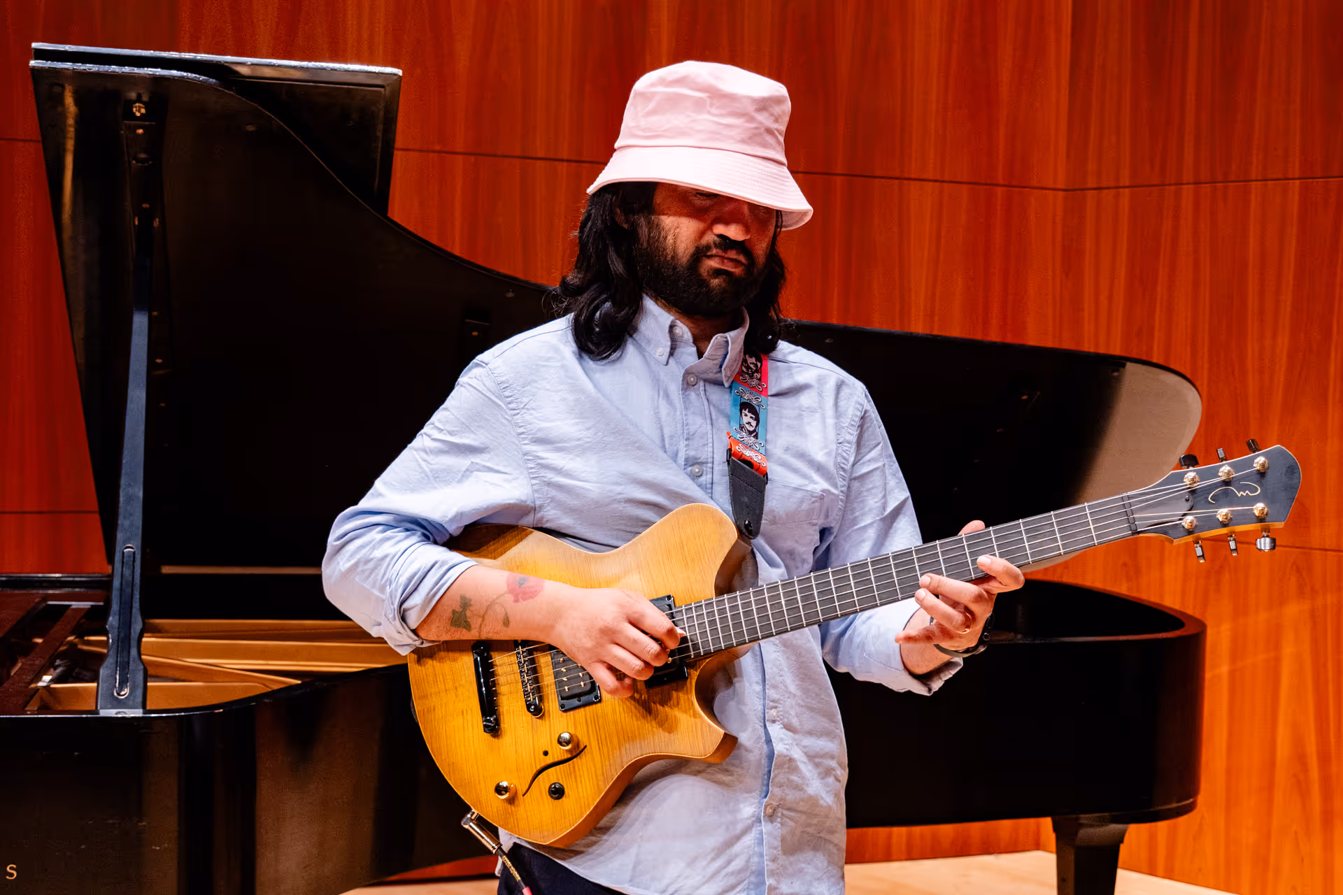 Eshaan Sood stands alone on stage, playing a bright yellow electric guitar in front of a grand piano. Wearing a light blue shirt and pink bucket hat, he is deeply focused on his performance, framed by the elegant wooden panels of the concert hall.