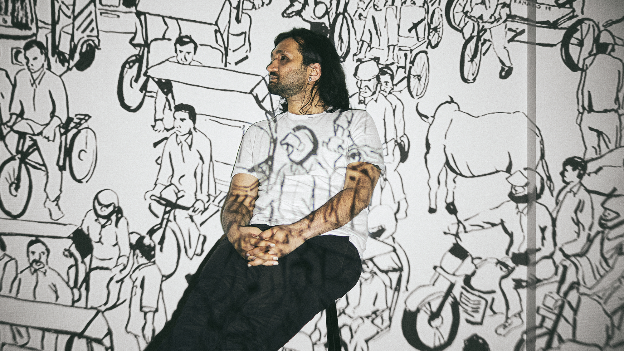 Eshaan Sood poses seated against a black-and-white projection of a chaotic Indian street scene, filled with sketched figures riding bicycles, walking, and navigating traffic. The visuals spill across his clothing and arms, blending him into the illustrated environment like a living mural.
