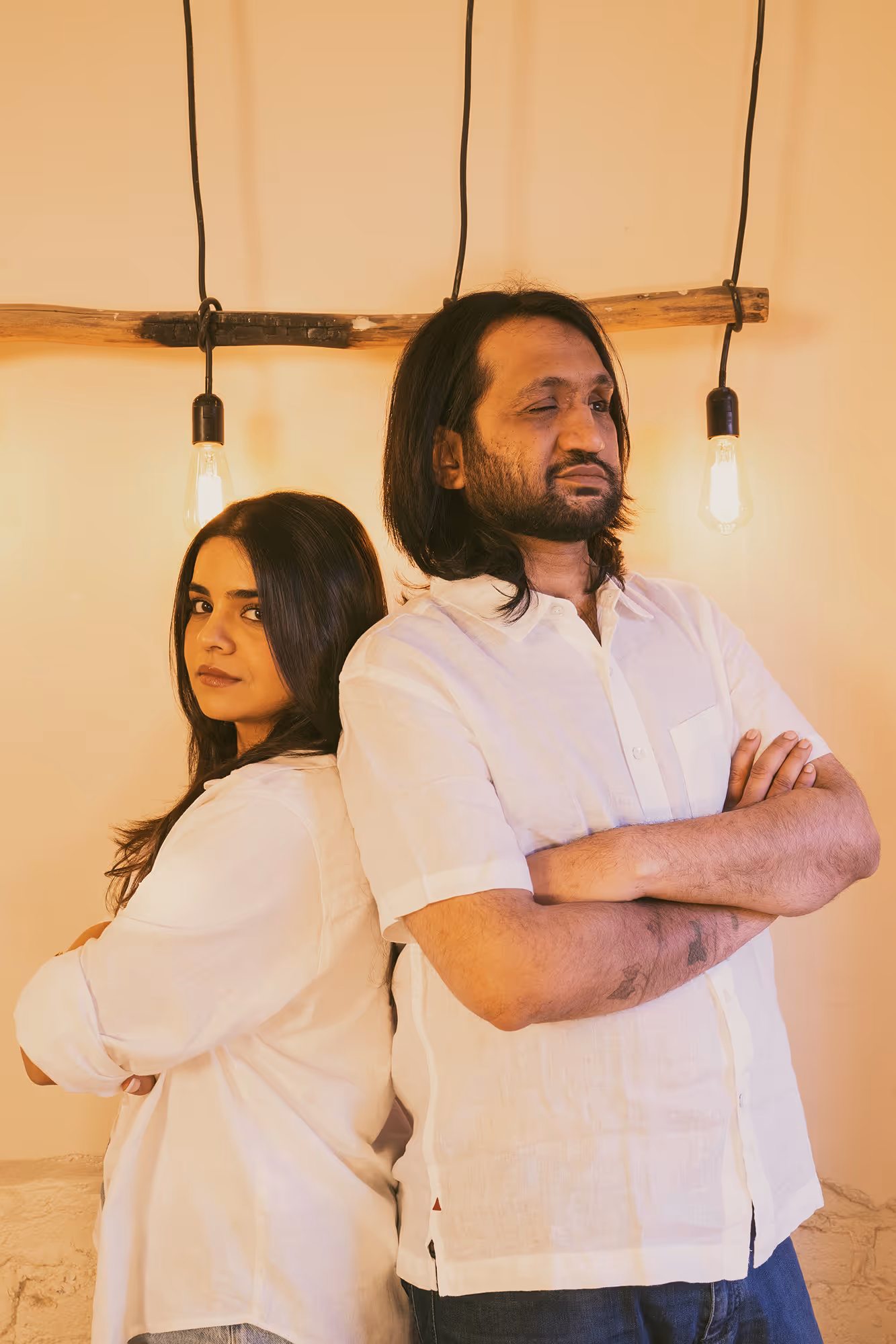 Eshaan Sood and Priyaana pose confidently with their arms crossed, standing back-to-back in a softly lit space. Both are dressed in white, and their body language is assertive yet calm. Edison bulbs hang above them, casting a glow that enhances the intimate atmosphere.