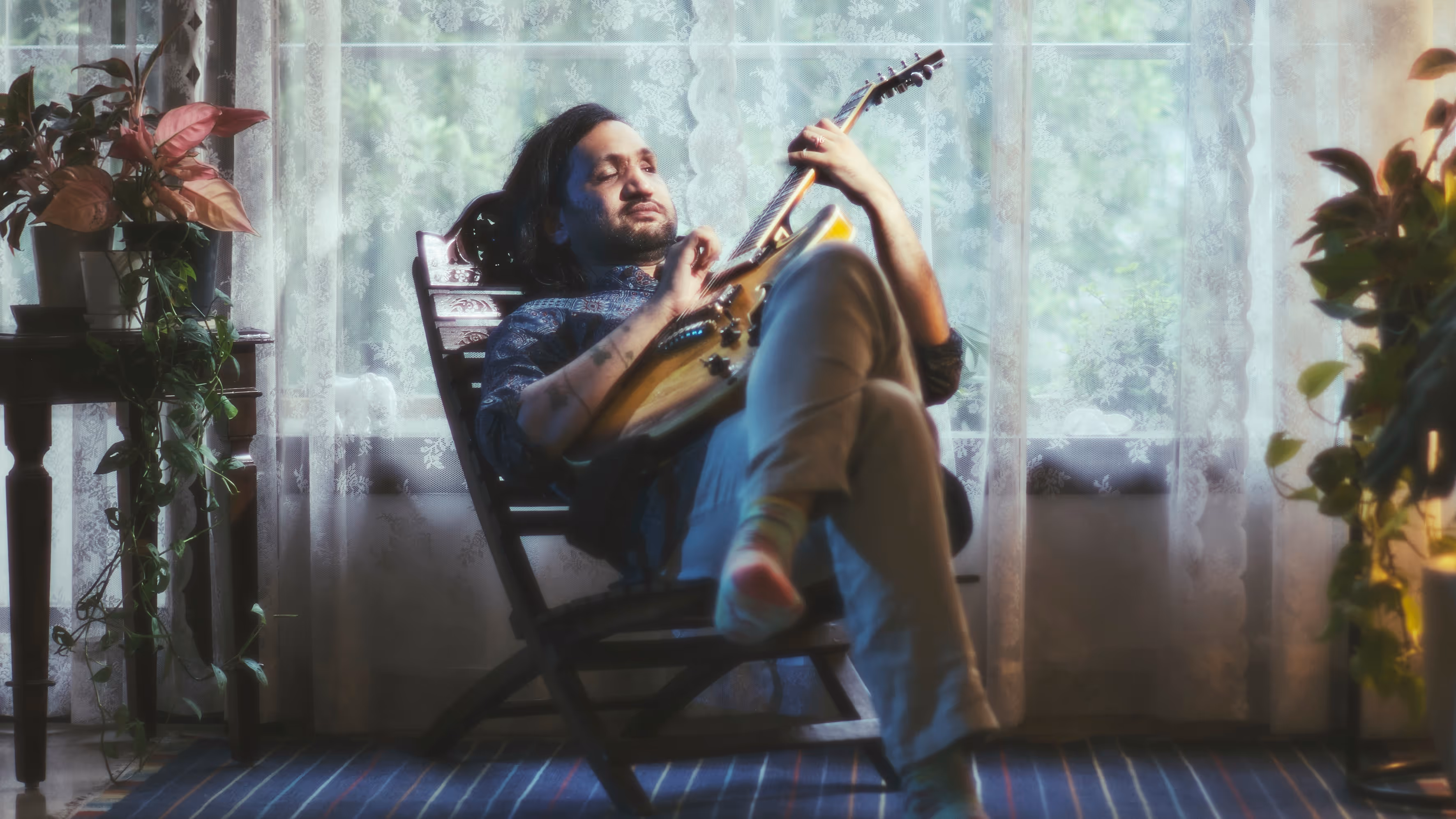 Eshaan Sood reclines with a guitar in hand, legs crossed, surrounded by houseplants on either side. The scene is soaked in warm, dreamlike light streaming through lace curtains. His expression is relaxed, absorbed in a moment of musical peace.
